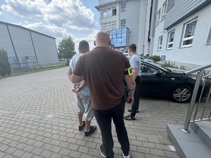 Zatrzymany mężczyzna stojący tyłem z kajdankami na rękach w obecności nieumundurowanych policjantów.