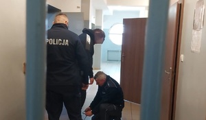 Pomieszczenie dla osób zatrzymanych. Dwóch policjantów umundurowanych zakłada zatrzymanemu kajdanki zespolone.