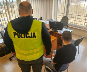 Pomieszczenie w którym są biurka i komputer. Po prawej stronie na krzesełku siedzi zatrzymany, a po lewej stronie koło niego stoi policjant w kamizelce z napisem Policja.