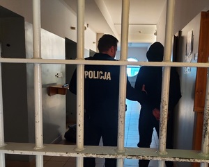 Za kratami tyłem do zdjęcia stoi policjant, na plecach ma napis Policja, po jego prawej stronie stoi zatrzymany, ma kaptur na głowie.
