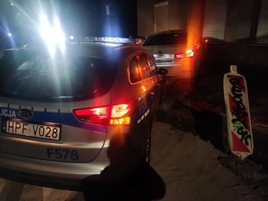Radiowóz stoi za samochodem, który stoi przed ścianą budynku. Jest noc, świeca się światła w samochodach.