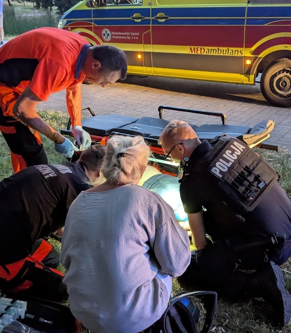Na trawniku leży na boku osoba, pochyla się nad nią policjant, dwóch ratowników medycznych i kobieta. W tle widać nosze i karetkę pogotowia.