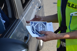Policjant trzyma w dłoni naklejki i ulotki kampanii "Mistrzu zwolnij". Policjant stoi obok samochodu.