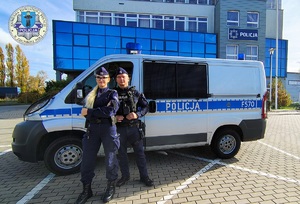 Umundurowani policjantka i policjant stoją przed radiowozem. W tle budynek komendy.