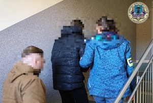 Policjanci prowadzą zatrzymanego po schodach budynku. Są wewnątrz budynku komendy.