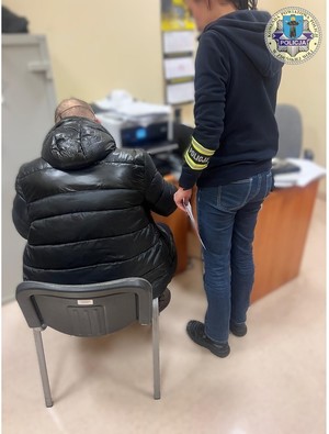 Zatrzymany siedzi tyłem do zdjęcia, podpisuje dokumenty, obok niego stoi policjant, ma na ramieniu opaskę.