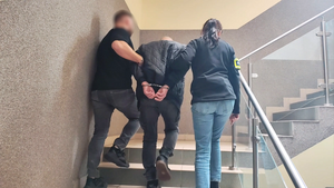 Policjanci prowadzą po schodach zatrzymanego mężczyznę, który ma założone kajdanki.