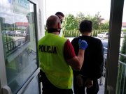 Policjanci  w kamizelkach odblaskowych z napisem policja, stoją tyłem, prowadzą zatrzymanego . Zatrzymany ubrany w czarny dres, ręce skute kajdankami, trzymane z tyłu. W tle parking policji.