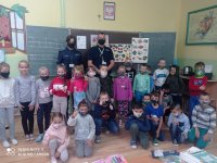 zdjęcie zrobione w sali lekcyjnej. Na tle tablicy stoją umundurowani policjanci przed nimi dzieci z kalsy 1 wszyscy maja nałożone maseczki ochronne. Ściany w klasie  ozdobione sa plakatami, rysunkami dzieci.