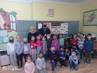 zdjęcie zrobione w sali lekcyjnej. Na tle tablicy stoj umundurowani policjanci nauczycielka  przed nimi dzieci z kalsy 1 wszyscy maja nałożone maseczki ochronne. Ściany w klasie  ozdobione sa plakatami, rysunkami dzieci.