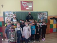 zdjęcie zrobione w sali lekcyjnej. Na tle tablicy stoją umundurowani policjanci przed nimi dzieci z kalsy 1 wszyscy maja nałożone maseczki ochronne. Ściany w klasie  ozdobione sa plakatami, rysunkami dzieci.