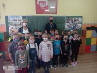 zdjęcie zrobione w sali lekcyjnej. Na tle tablicy stoją umundurowani policjanci przed nimi dzieci z kalsy 1 wszyscy maja nałożone maseczki ochronne. Ściany w klasie  ozdobione sa plakatami, rysunkami dzieci.
