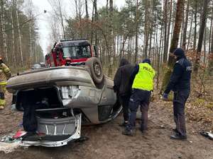 przewrócony samochód do góry kołami, widoczni są policjanci i strażacy, policyjny radiowóz, wóz strażacki.