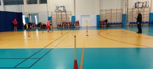 policjant w mundurze, dziecko biegnie przez salę gimnastyczną. na ławkach siedzą uczniowie.