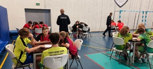 policjant w mundurze, dzieci siedzą przy stolikach, w kolorowych ubraniach.