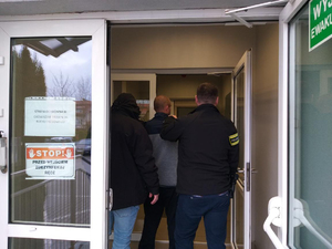dwóch nieumundurowanych policjantów prowadzi mężczyznę, który ma kajdanki założone na ręce trzymane z tyłu. Wchodzą do budynku.