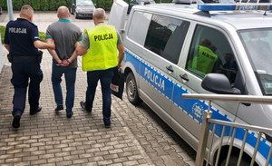 mężczyzna w kajdankach prowadzony przez dwóch policjantów, obok radiowóz policyjny.