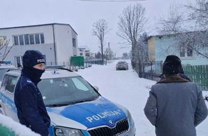 radiowóz, policjant, osoba w kurtce i czpace, budynki.