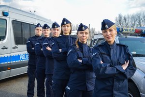 Zdjęcie przedstawia sześciu funkcjonariuszy ubranych w mundury ćwiczebne, stojących w rzędzie jedno za drugim.
Po prawej i lewej stronie znajdują się radiowozy.