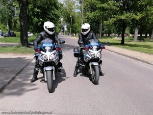 Obraz przedstawia dwóch policjantów ruchu drogowego siedzących na motocyklach oznakowanych, w kaskach i mundurach motocyklowych, patrzących się na siebie. Stoją oni na drodze