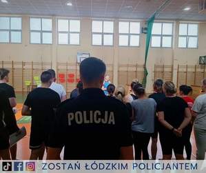 Policjant stojący na tle uczestników próbnego testu sprawności fizycznej, odbywającego się na terenie Oddziału Prewencji Policji w Łodzi.